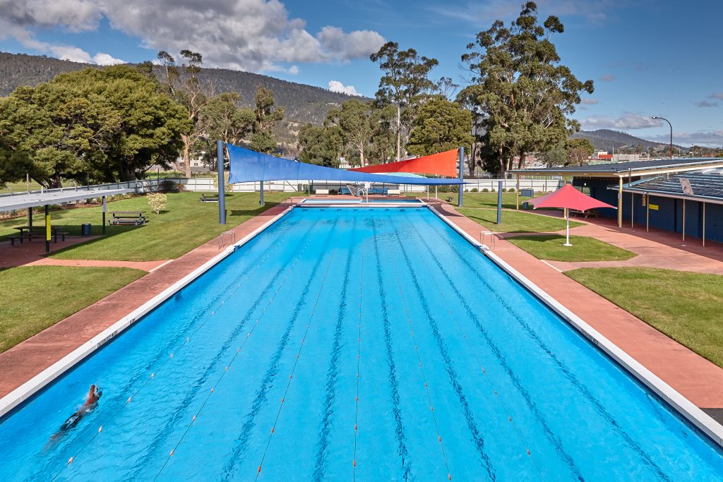 A long outdoor swimming pool with lane dividers is surrounded by grass and trees. A swimmer is in one lane. Red and blue shade sails are overhead. The sky is partly cloudy, and mountains are visible in the background.