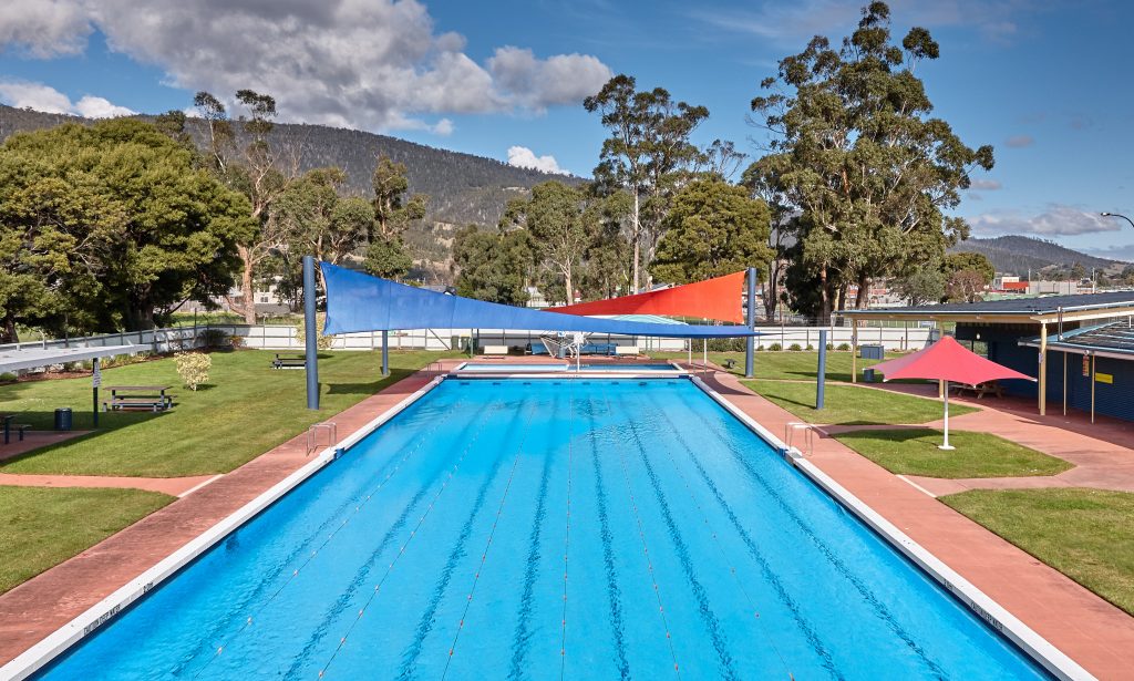 Outdoor swimming pool with clear blue water, surrounded by green lawns and trees. Two colorful shade sails, one blue and one red, stretch over one end of the pool. Hills and blue sky with clouds are in the background.