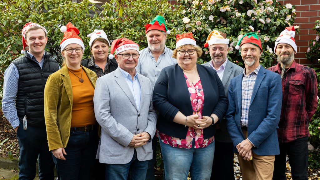 A group of nine people, wearing festive hats, smile and pose for a photo outdoors. They are standing in front of green foliage and a brick wall. The scene conveys a cheerful, holiday atmosphere.