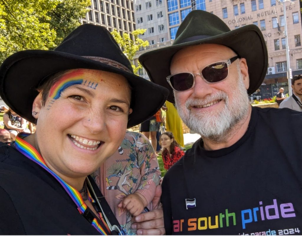 Two people wearing black hats smile at the camera during an outdoor event. The person on the left has a rainbow painted on their cheek, and both wear clothing with pride-themed accessories. Buildings and trees are visible in the background.