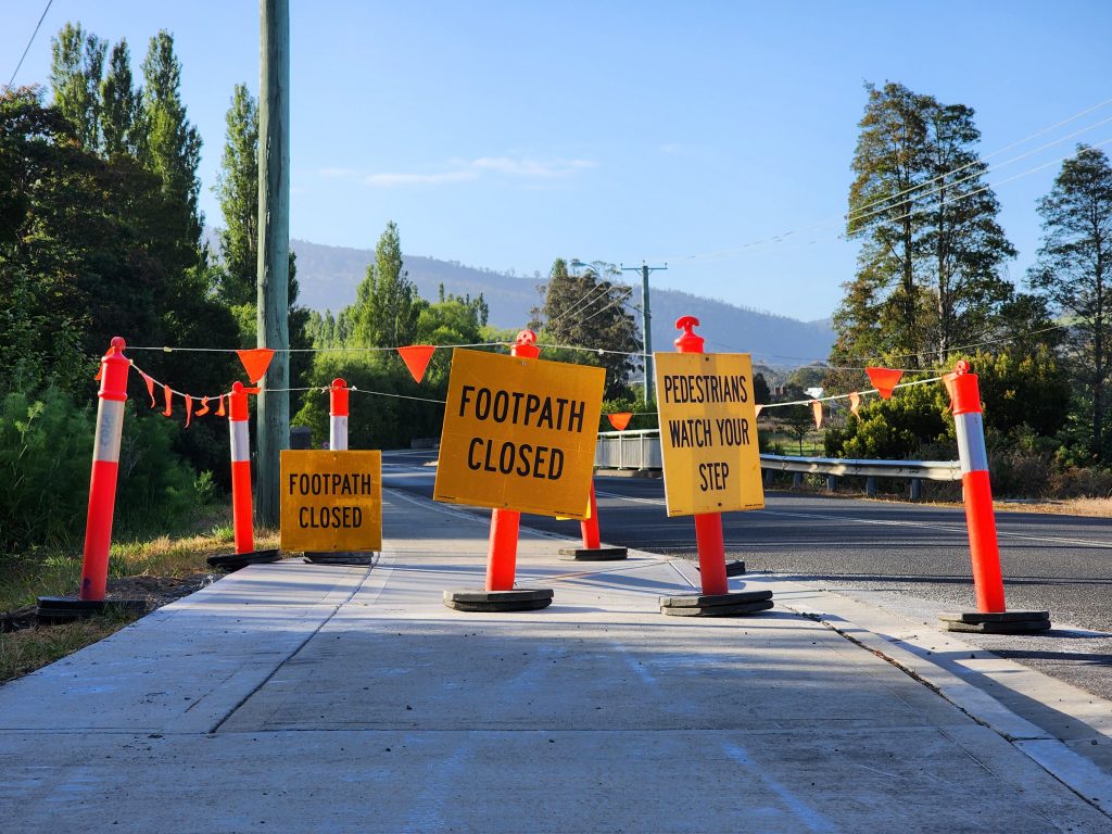 Sidewalk under construction with Footpath Closed and Pedestrians Watch Your Step signs. Orange cones and barriers redirect pedestrians. Background features lush greenery and distant hills.