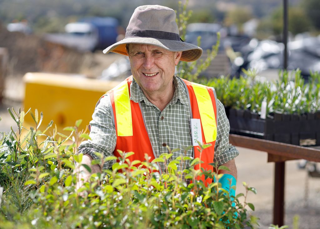 A man in a plaid shirt and wide-brimmed hat stands among green plants, wearing a fluorescent safety vest labeled with a name tag. He smiles while tending to the plants outdoors.