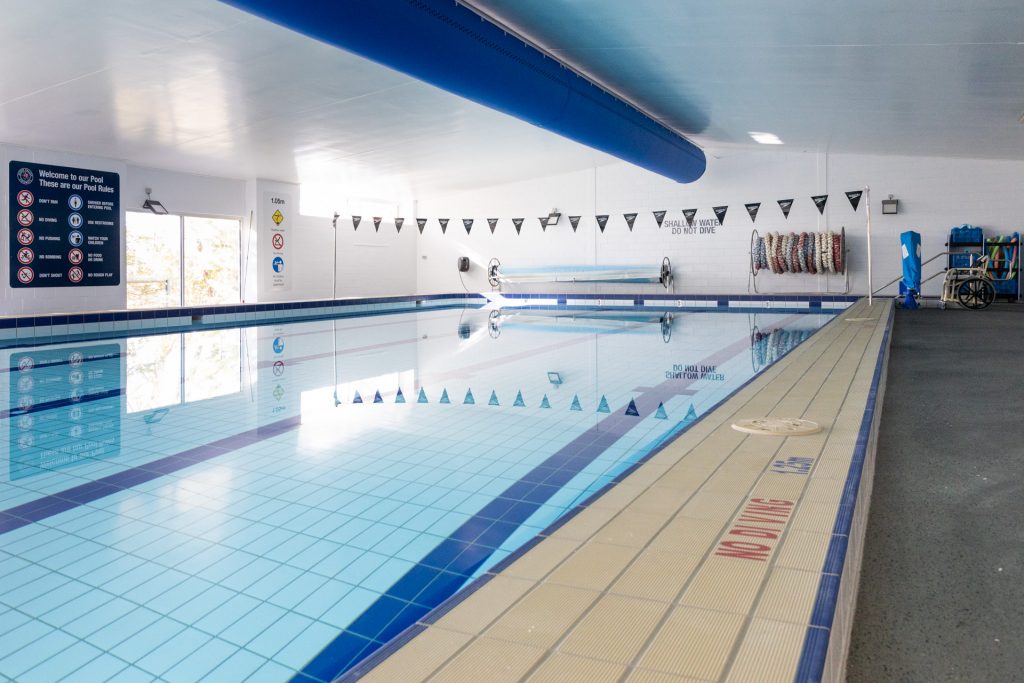 Indoor swimming pool with clear water and tiled floor. Safety signs and swimming lane dividers are visible. A large blue ventilation duct runs along the ceiling, and a stack of pool noodles is on the far wall next to a wheelchair-accessible lift.