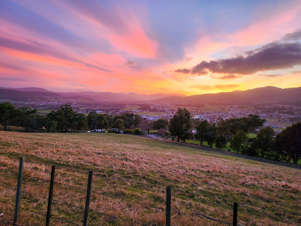 Sunset view of a peaceful rural landscape with a grassy field in the foreground and a fence. Houses and trees fill the midground, backed by rolling hills. The sky is painted in vibrant pink, orange, and purple hues.