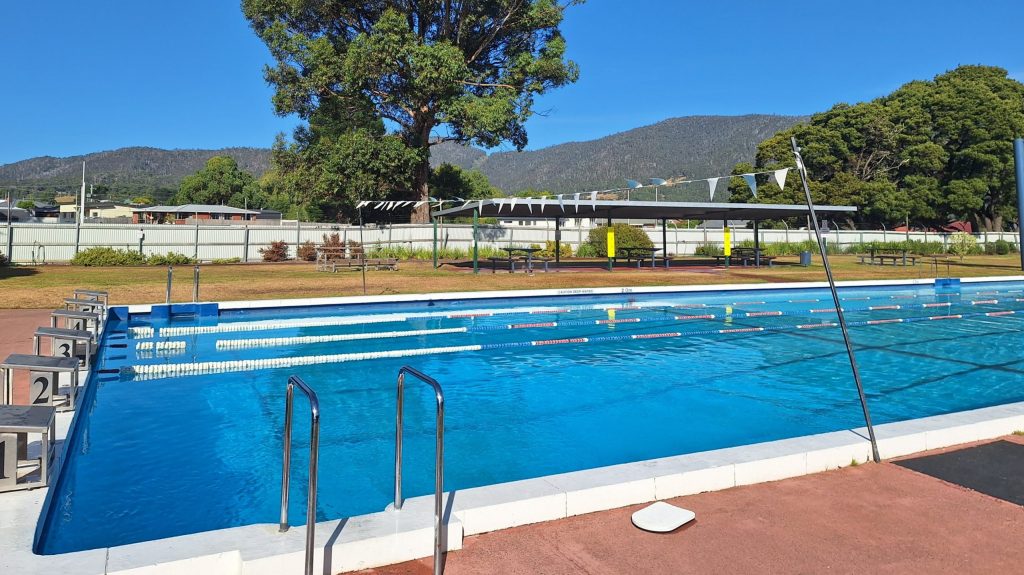 Outdoor swimming pool with numbered lanes under a clear blue sky. Tall trees and mountains are visible in the background. Pool is empty and surrounded by a fence.