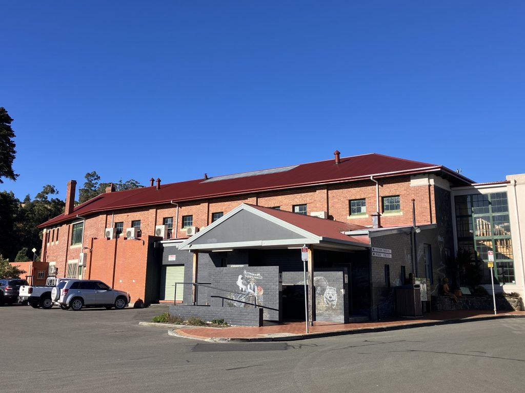 Brick building with a red roof and a smaller gray annex in front. Cars are parked on the left, and a blue sky above. Theres a sidewalk leading to a ramp and stairs. Trees are partially visible in the background.