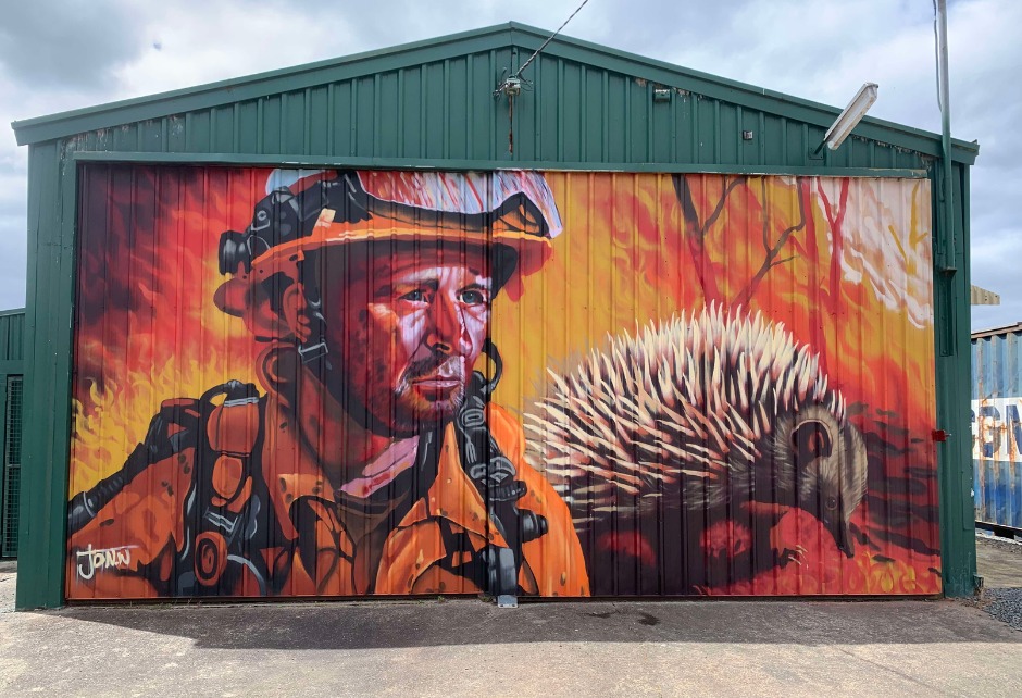 A mural on a green metal building depicts a firefighter in orange gear and helmet beside an echidna, with bright flames and smoke in the background, highlighting a wildfire scene.