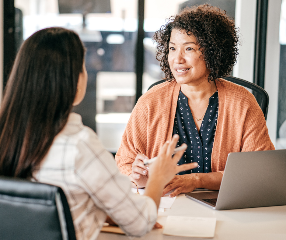 Two women sit across a desk from each other in an office, engaged in a conversation. One woman is holding a pen and notepad, while the other listens attentively with a laptop open in front of her.