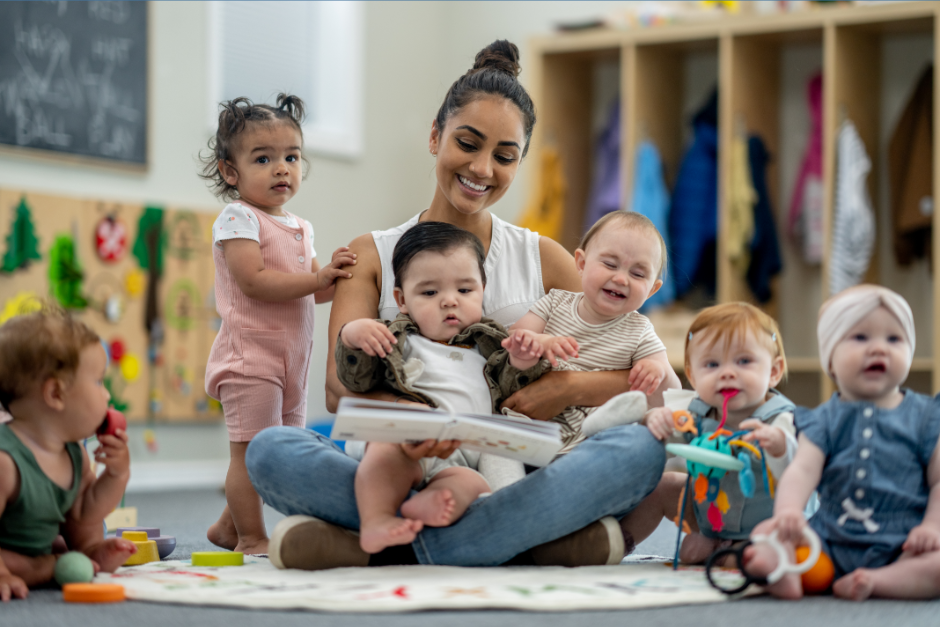 A woman sits on the floor reading a book to a group of six babies and toddlers who are gathered around her, some sitting and some crawling, in a colorful classroom or daycare setting.
