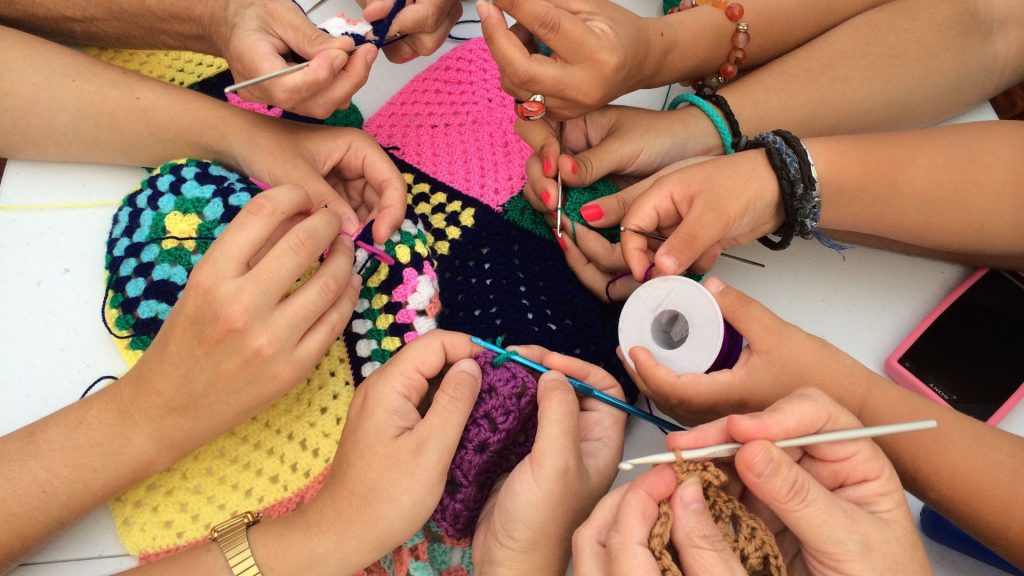 A group of diverse hands crocheting together, creating a colorful patchwork of yarn. Crocheting tools and various yarn colors are visible, with vibrant squares and patterns forming at the center of their collaborative effort.
