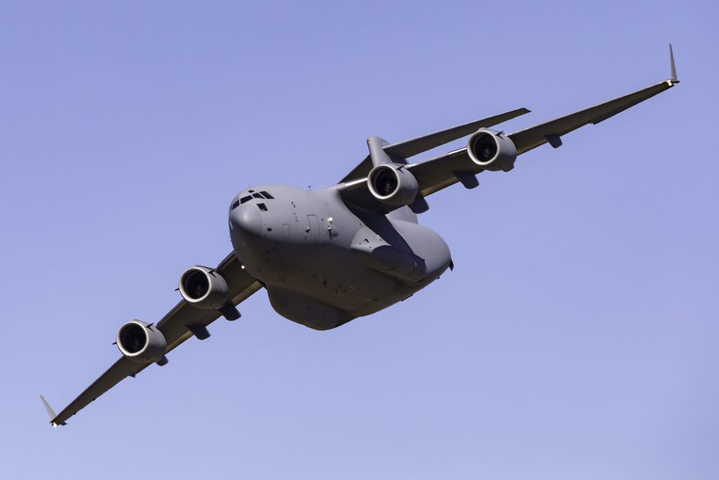 A large gray military cargo aircraft with four engines flies at a low angle against a clear blue sky.