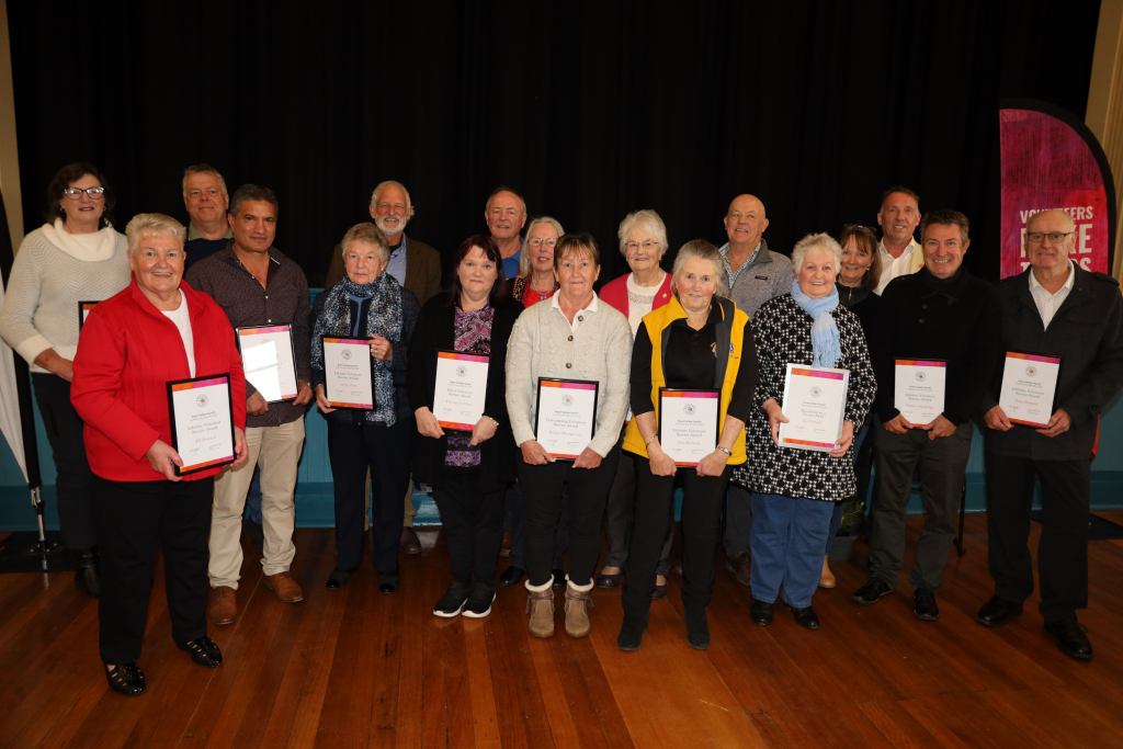 A group of people stand indoors in two rows, smiling and holding certificates. They are positioned on a wooden floor in front of a black curtain, with a banner partly visible on the right.