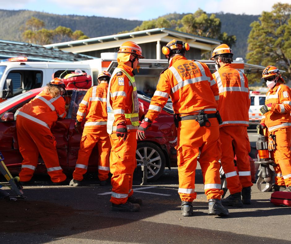 A group of emergency rescue workers in orange uniforms assist at the scene of a car accident, gathered around a damaged red car, with rescue equipment in use and a building in the background.
