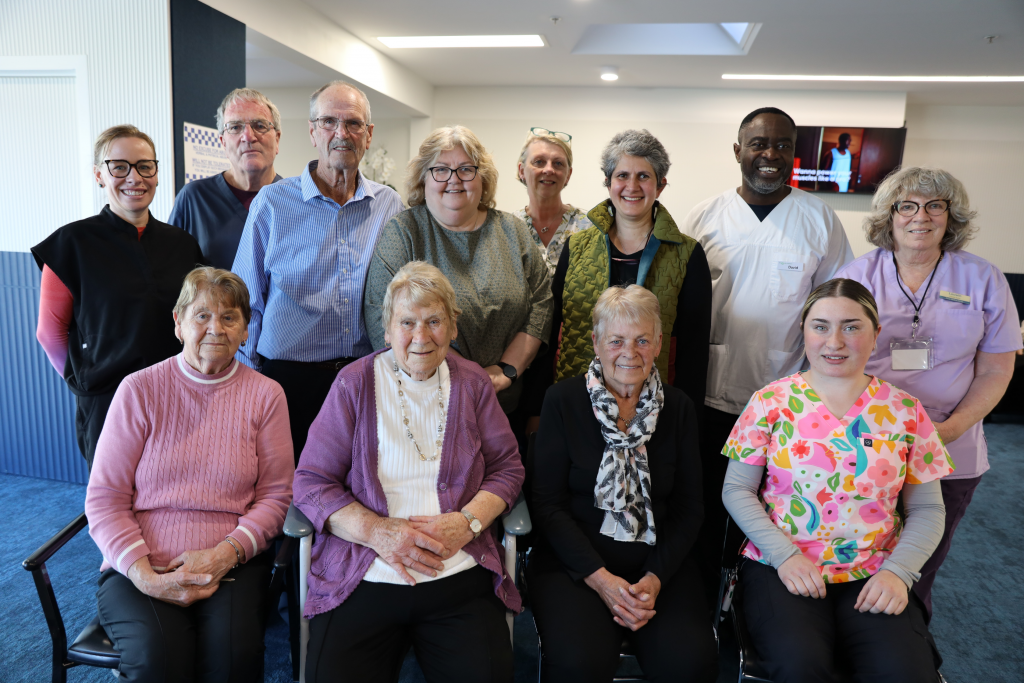 A group of thirteen people, including elderly women seated in front and healthcare staff, pose together indoors in a well-lit room, smiling for the camera.