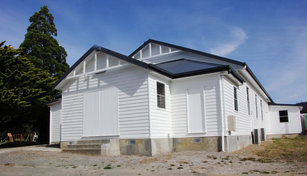 A white wooden building with a dark metal roof sits on a concrete foundation, surrounded by gravel and some grass, under a blue sky with wispy clouds and trees in the background.