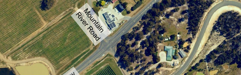 Aerial view of a rural intersection with labeled roads: Mountain River Road and a partially visible road. Surrounding areas include fields, trees, buildings, and a curved dirt track.