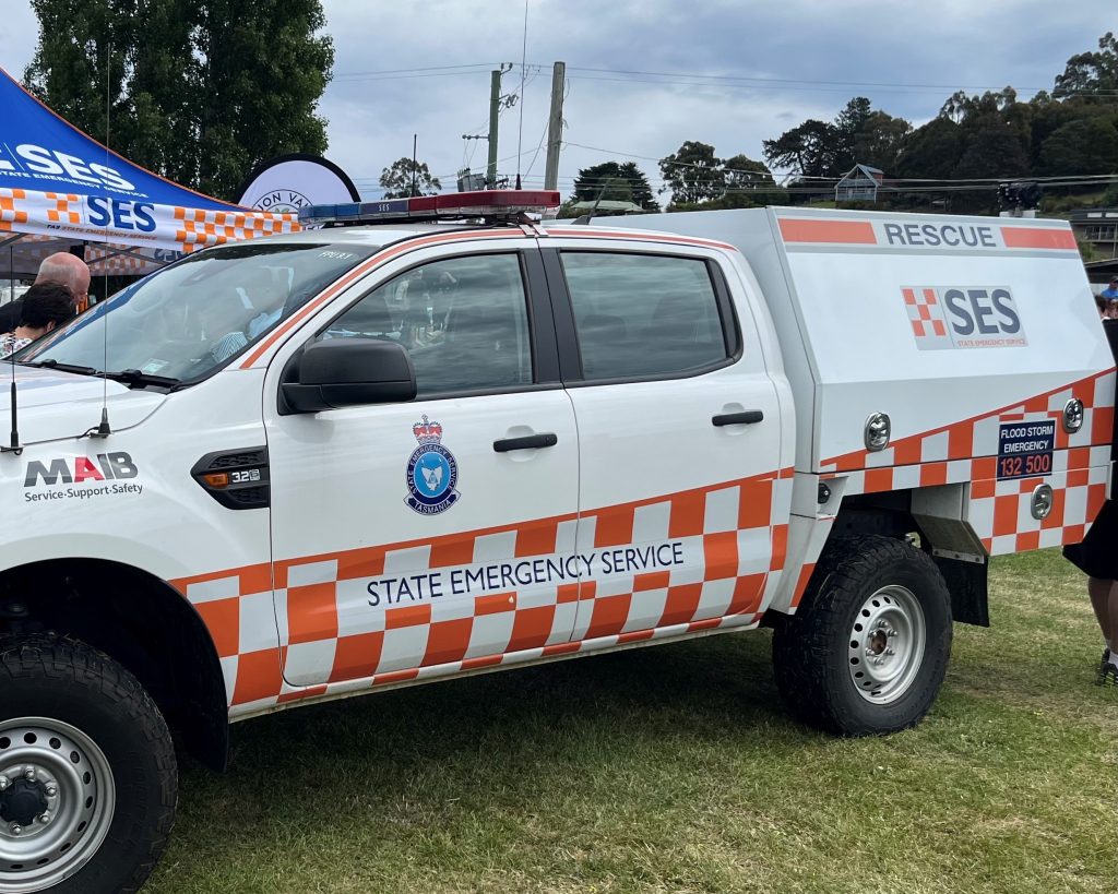 A white State Emergency Service (SES) rescue vehicle with orange checkerboard markings is parked on grass at an outdoor event. Emergency contact information and SES logos are visible on the side.