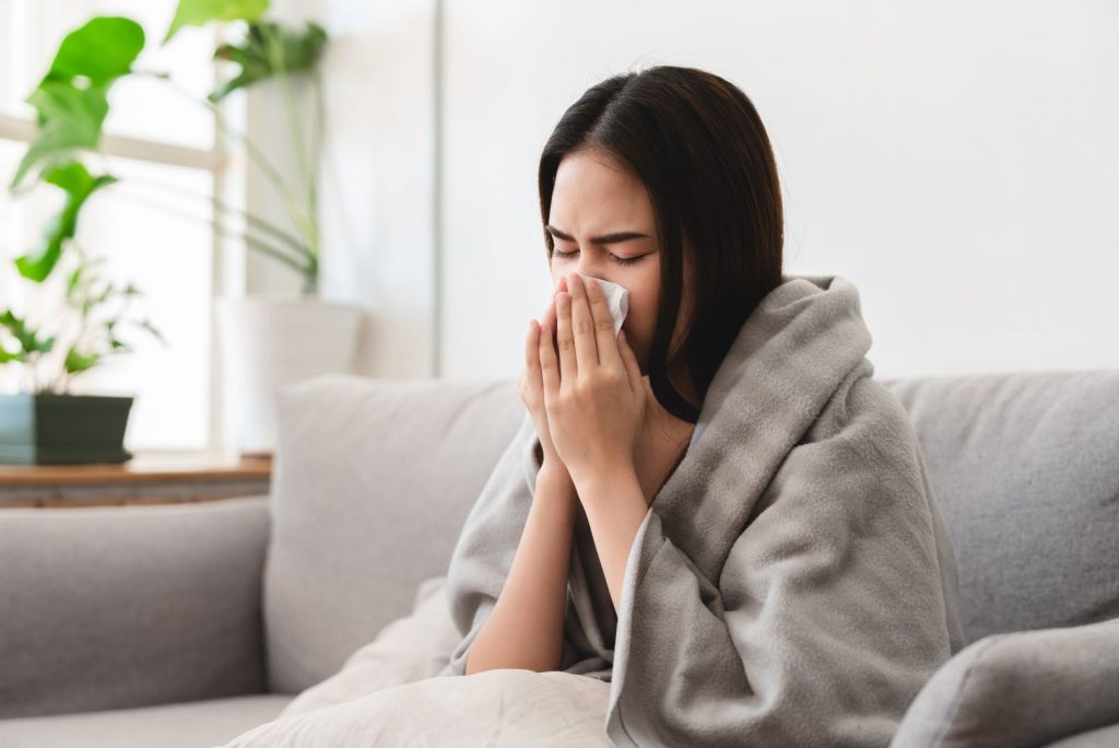 A young woman wrapped in a gray blanket sits on a couch, holding a tissue to her nose and appearing to sneeze or blow her nose; plants and a window are in the background.