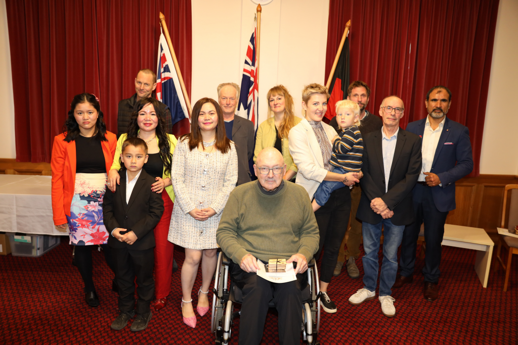 A group of people, including men, women, and children, pose indoors in front of three flags and red curtains. One man in a wheelchair sits in front, while others stand behind him, dressed in semi-formal attire.