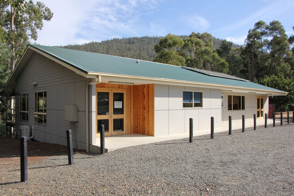 A modern, single-story building with a green roof and beige siding, surrounded by trees and mountains, with a gravel parking area and black bollards in front of the entrance.