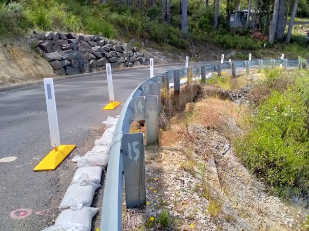 A curved road with a metal guardrail and orange safety barriers, bordered by sandbags. The right side shows eroded ground and a steep drop-off with rocks and vegetation. Trees and bushes line the roadside.