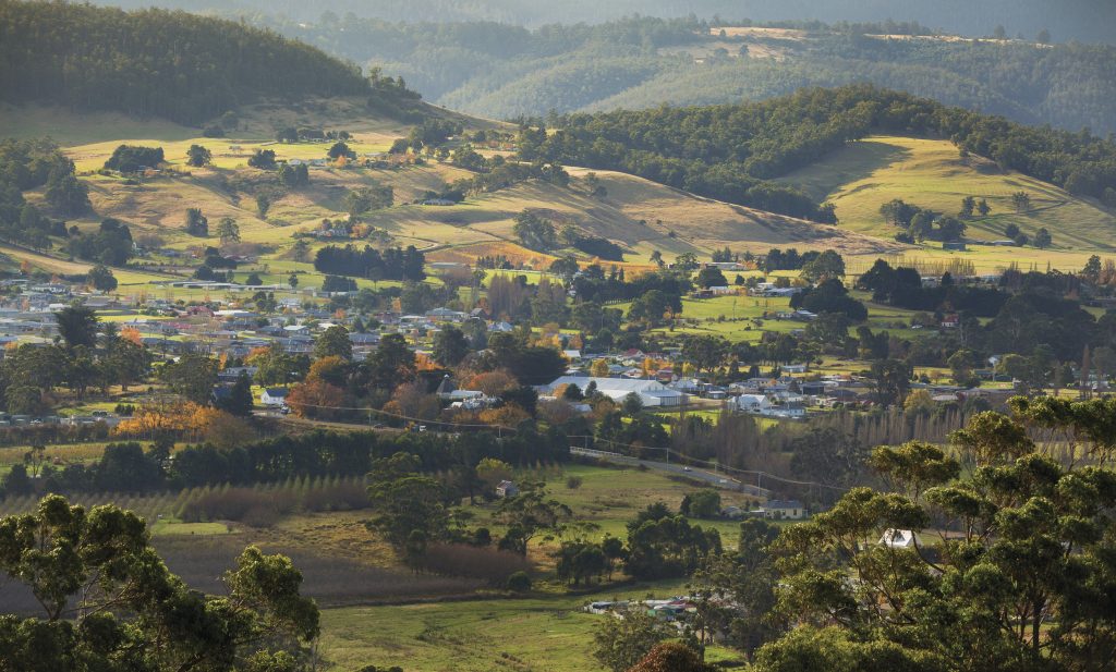 A small rural town stretches across a green valley, surrounded by rolling hills and scattered trees under soft sunlight, with houses, open fields, and forested slopes visible in the landscape.