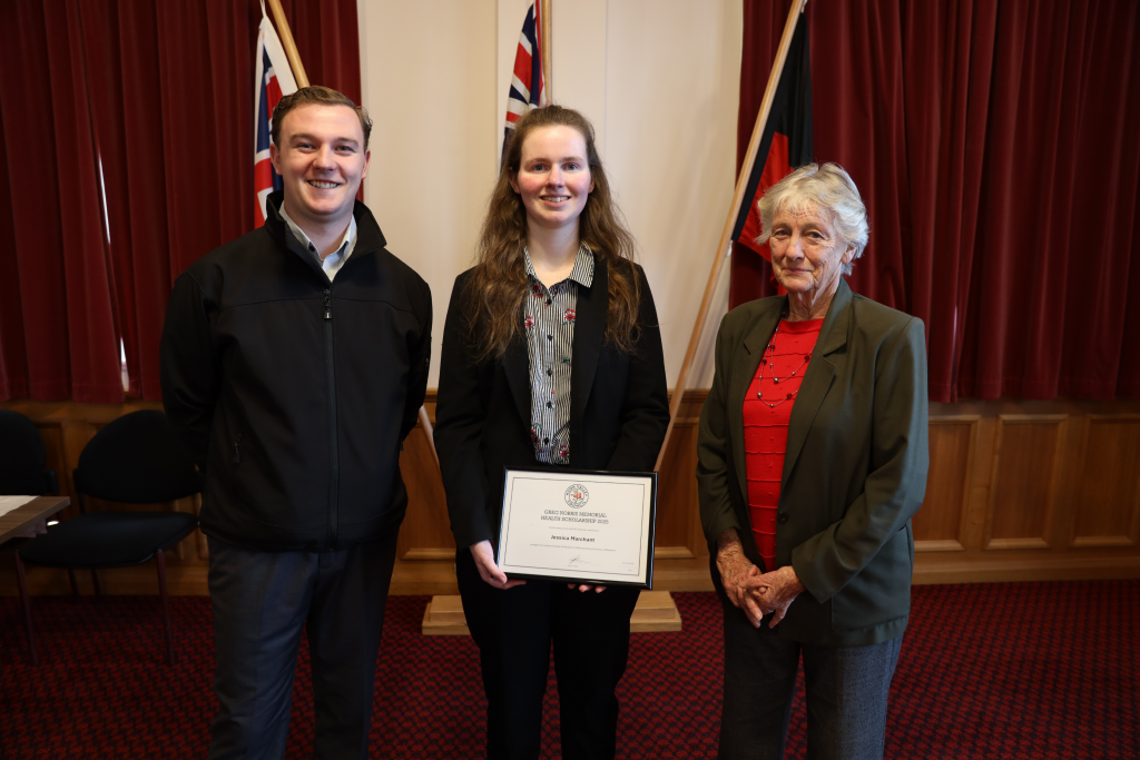 Three people stand indoors in front of red curtains and two flags. The person in the center holds a certificate and smiles, with a person on each side also smiling at the camera.