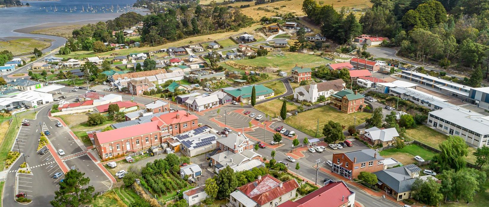 Aerial view of a small town with red-roofed buildings, green fields, trees, winding roads, and a river with moored boats in the background, under a blue sky with light clouds.