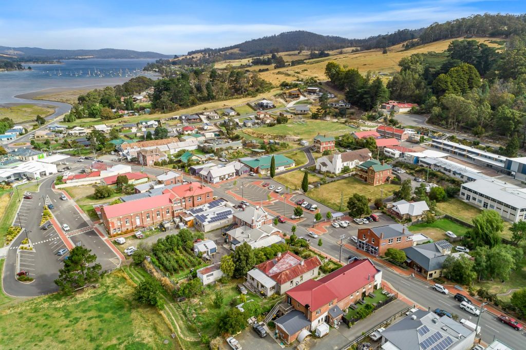 Aerial view of a small town with red-roofed buildings, green trees, winding roads, and a river with boats in the background, surrounded by rolling hills and open fields under a blue sky.