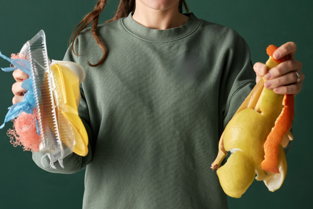 A person in a green sweatshirt holds plastic waste in one hand and fruit peels in the other, standing against a green background. Their face is partially out of frame.
