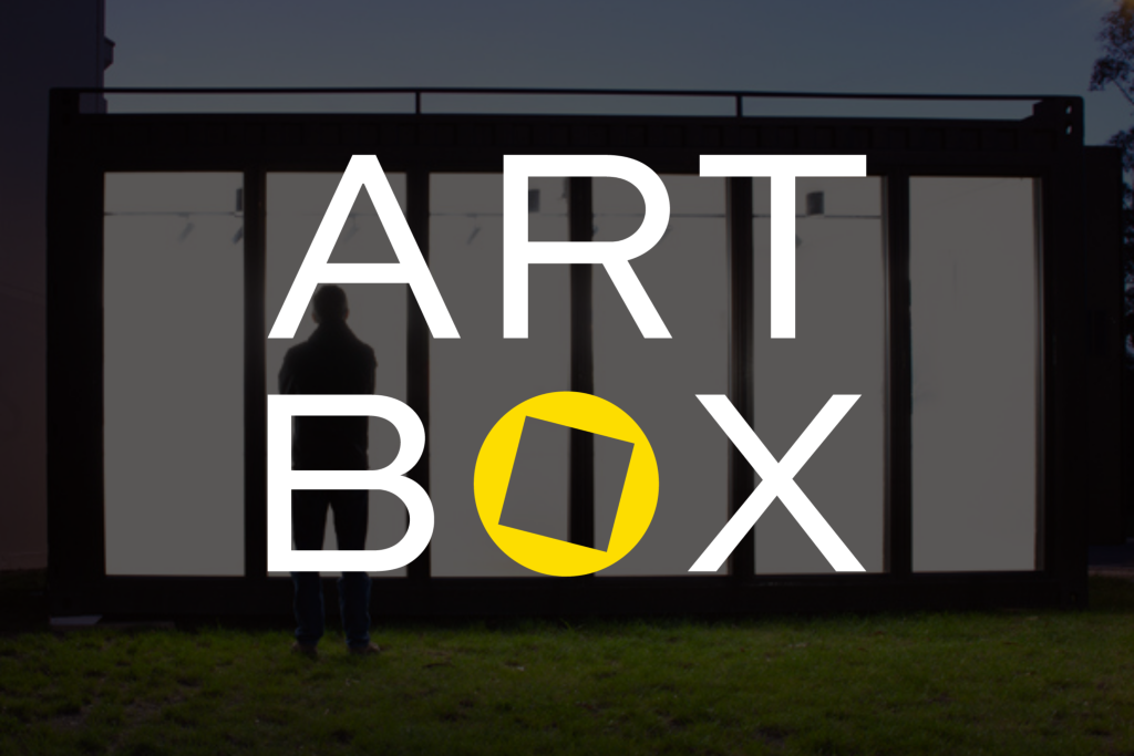 A person stands in front of a large glass display case at dusk. Overlaid text reads ART BOX, with the letter O replaced by a tilted yellow square inside a circle.