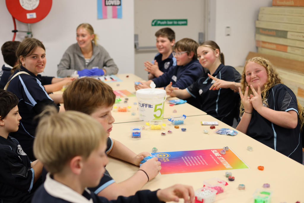 A group of school children sit around a table working on crafts, smiling and interacting. A teacher sits at the end of the table. Colorful beads and posters are spread out on the table.
