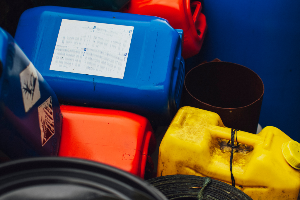 A collection of colorful plastic containers, including blue, yellow, and red jerry cans, some with labels and warning symbols, stacked closely together with a black hose visible in the foreground.