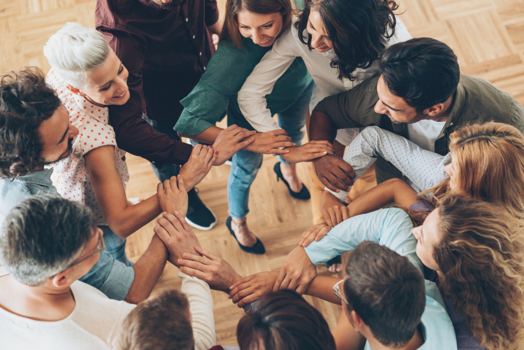 A diverse group of people stands in a circle with their hands joined in the center, smiling and appearing united and enthusiastic, shown from an overhead perspective on a wooden floor.