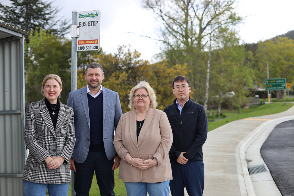 Four adults stand smiling near a Tassielink bus stop on a suburban street, with greenery and road signs in the background. Three people wear jackets and one wears a blazer. It is daytime.