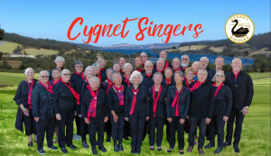 A choir group of about 30 adults stands together outdoors on grass, wearing black outfits with red scarves. Behind them is a scenic landscape with hills, trees, and water. Text reads “Cygnet Singers.”.
