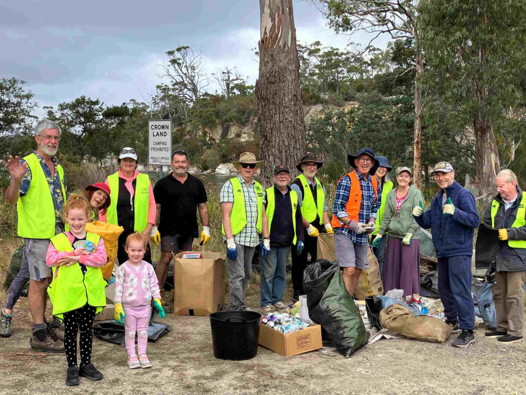 A group of people, including children, wearing yellow safety vests pose together outdoors with collected trash bags and recycling. Trees and a sign are visible in the background; everyone appears happy and proud.