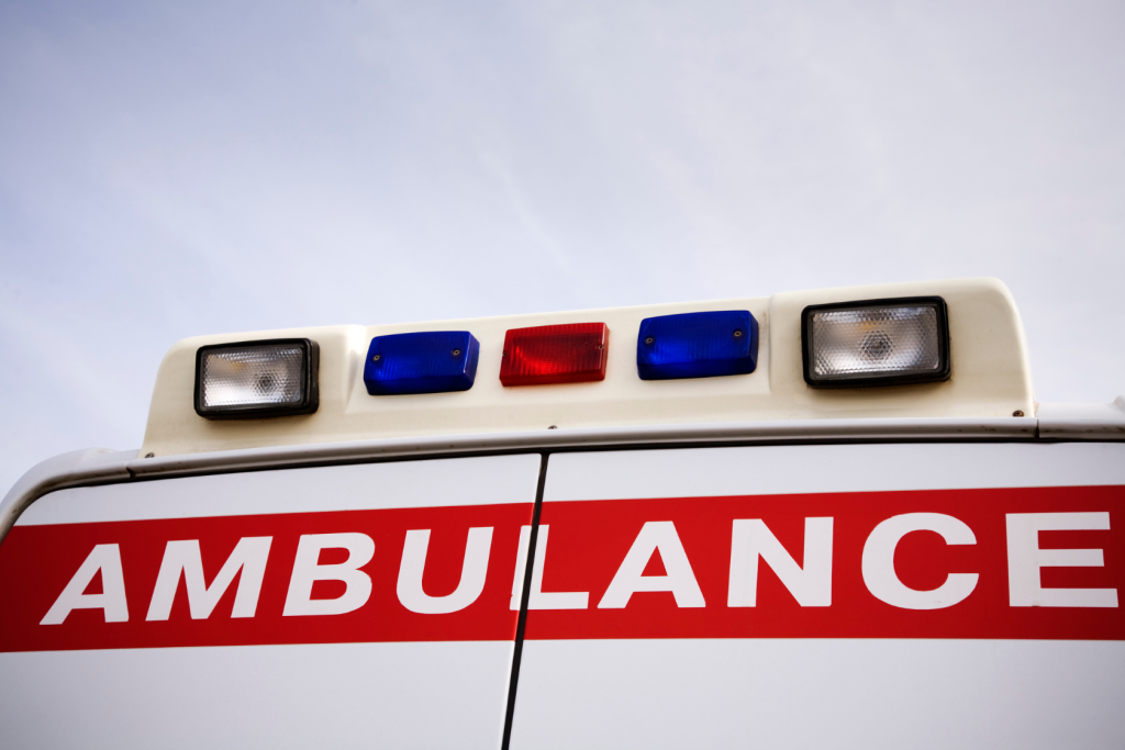 The image shows the back of an ambulance with AMBULANCE written in bold red letters and emergency lights on top against a cloudy sky.