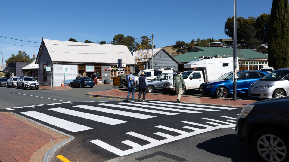 Three people cross a wide, freshly painted crosswalk on a sunny day in front of small shops and parked cars in a quiet town. The sky is clear and blue, and the area appears calm and tidy.