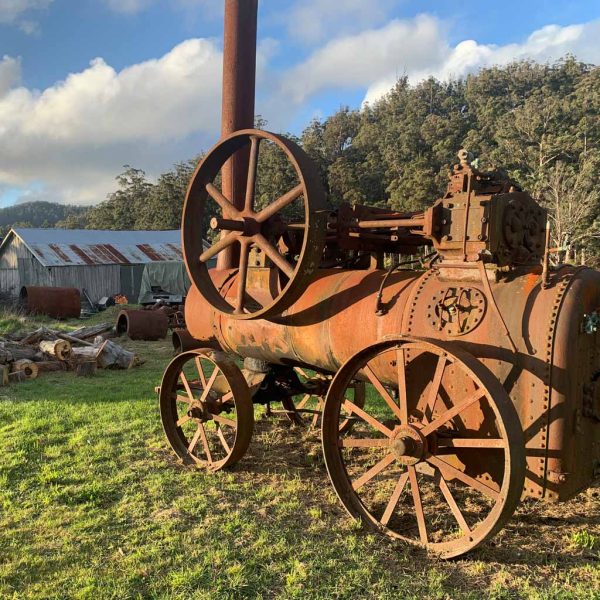 A large, rusted steam engine with metal wheels stands on grass near old machinery and a weathered shed, with trees and a partly cloudy sky in the background.