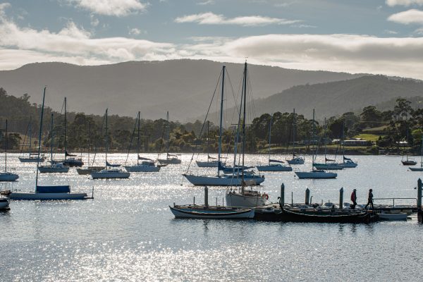 Several sailboats and yachts are anchored on a sparkling, sunlit body of water with mountains and trees in the background. A few people stand on a dock in the foreground under a partly cloudy sky.