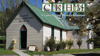 A cream-colored building with a green roof houses the Cygnet Living History Museum. A sign outside reads Proudly Open, and trees with white blossoms frame the scene. The museum logo and name appear at the top.