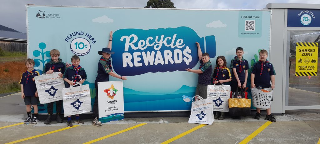 A group of Scouts in uniform stand in front of a “Recycle Rewards” container, holding bags and bins with recycling. A blue and white sign promotes a 10-cent refund for recycling.