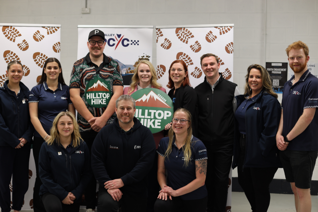 A group of twelve people pose indoors, some standing and some kneeling, in front of banners with the PCYC logo and a Hilltop Hike sign. They are smiling and wearing navy blue uniforms or casual clothing.
