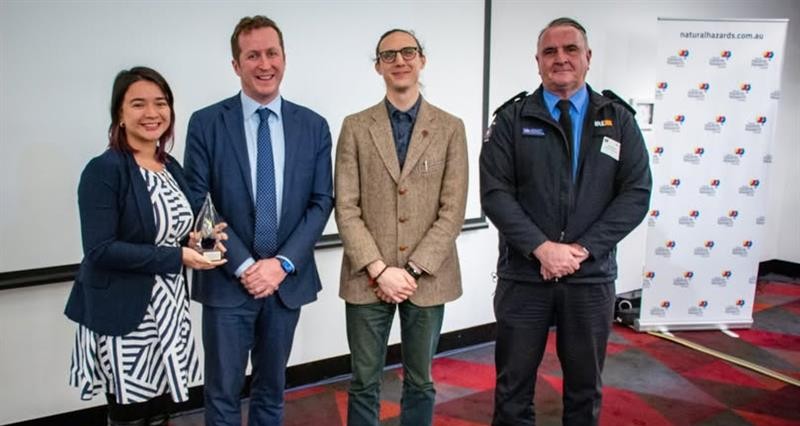 Four people stand side by side indoors at an event; the woman on the left holds a trophy. Behind them is a pull-up banner with logos and a projection screen. The group is smiling, dressed in business and uniform attire.