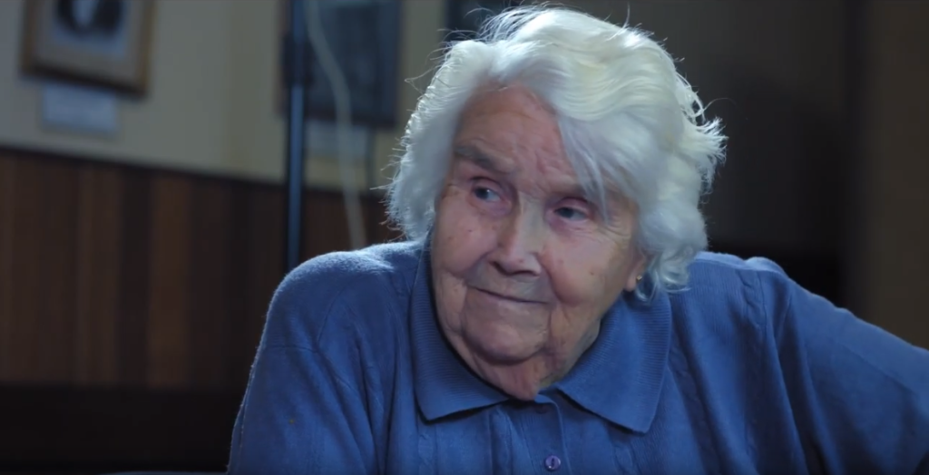An elderly woman with white hair and a blue collared shirt sits indoors, looking thoughtfully to the side. The background is softly lit with wooden paneling and framed pictures on the wall.