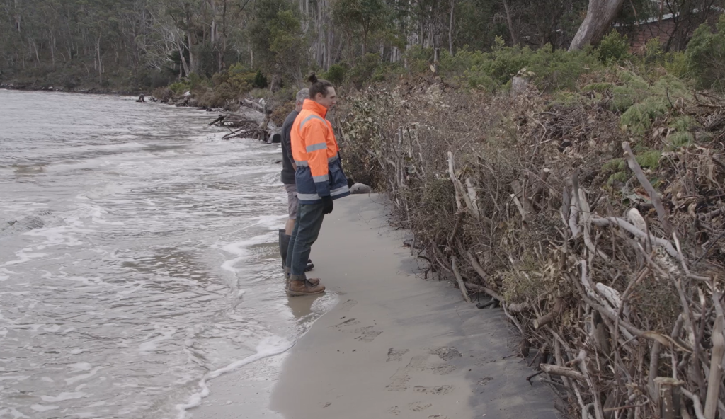 A person in an orange high-visibility jacket and boots stands on a narrow, sandy shoreline, observing eroded vegetation and debris along the edge of the water, with trees in the background.