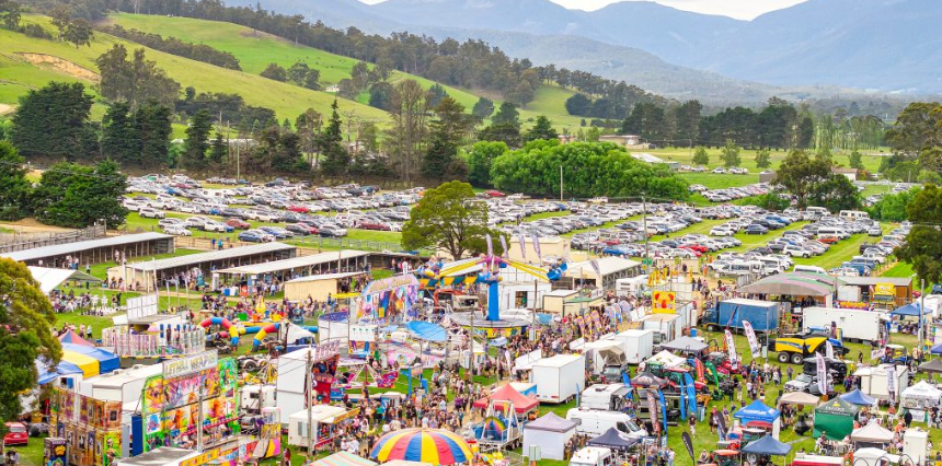 Aerial view of a lively outdoor fair with colorful tents, food stalls, amusement rides, crowds of people, and a parking lot full of cars, set against green hills and distant mountains.