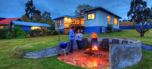 Two people stand by a fire pit in a circular patio area, surrounded by chairs, with a blue house and green lawn in the background during dusk.