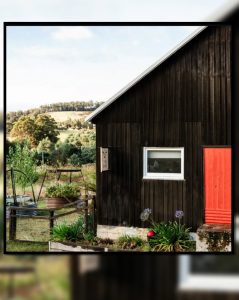 A rustic black wooden building with a bright red door and white-framed window stands in a garden, surrounded by greenery and potted plants, with fields and trees in the background.